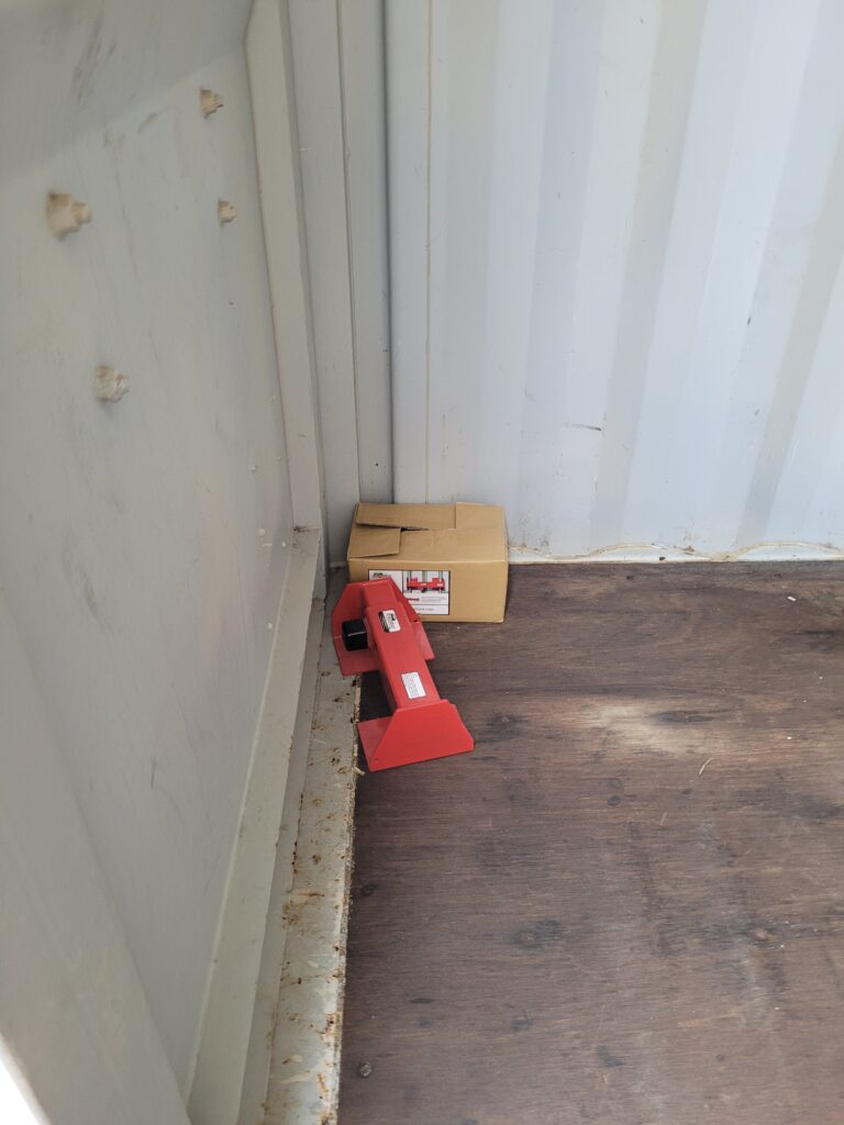 the interior of a 20ft storage container. The walls are made of corrugated metal. The floor is wooden, showing signs of use with stains and discoloration. Inside, there is a red piece of equipment (possibly a tool or machinery part) leaning against the wall, along with a cardboard box placed nearby.