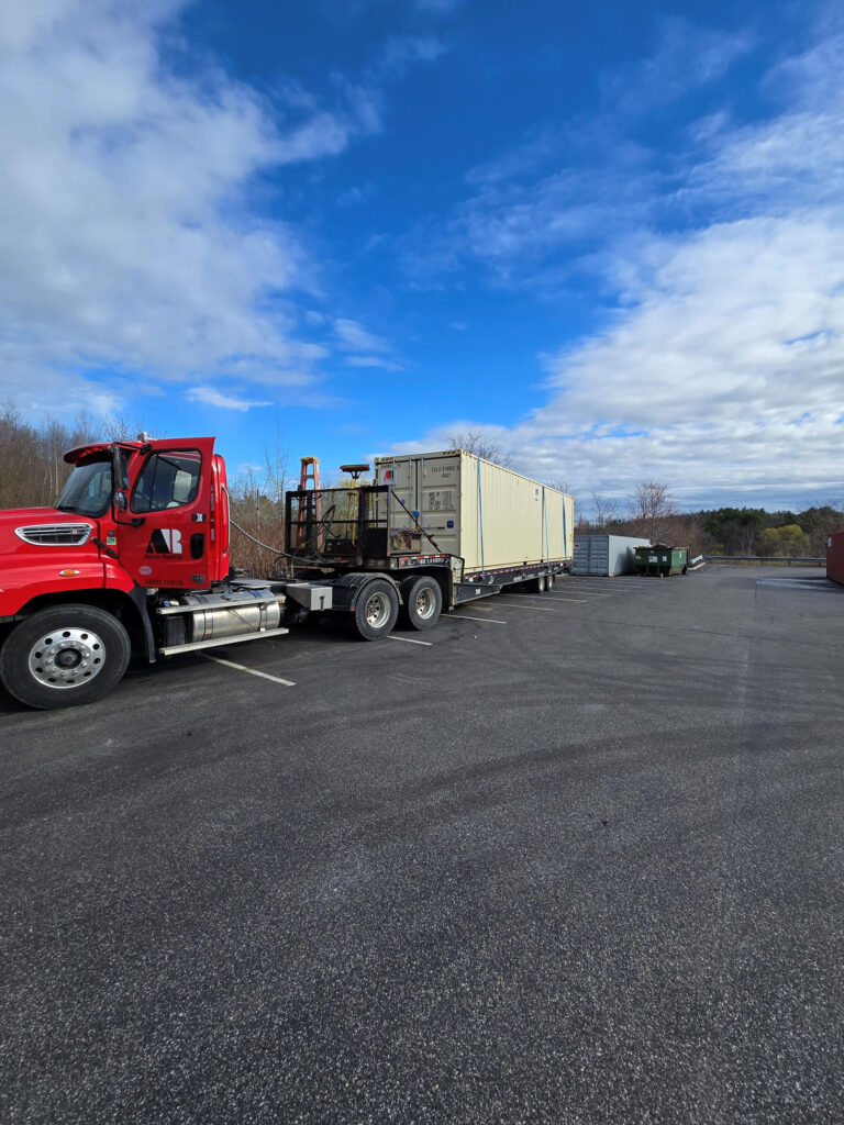 Red semi-truck with a beige shipping container on a flatbed trailer in a parking lot under a blue, partly cloudy sky.