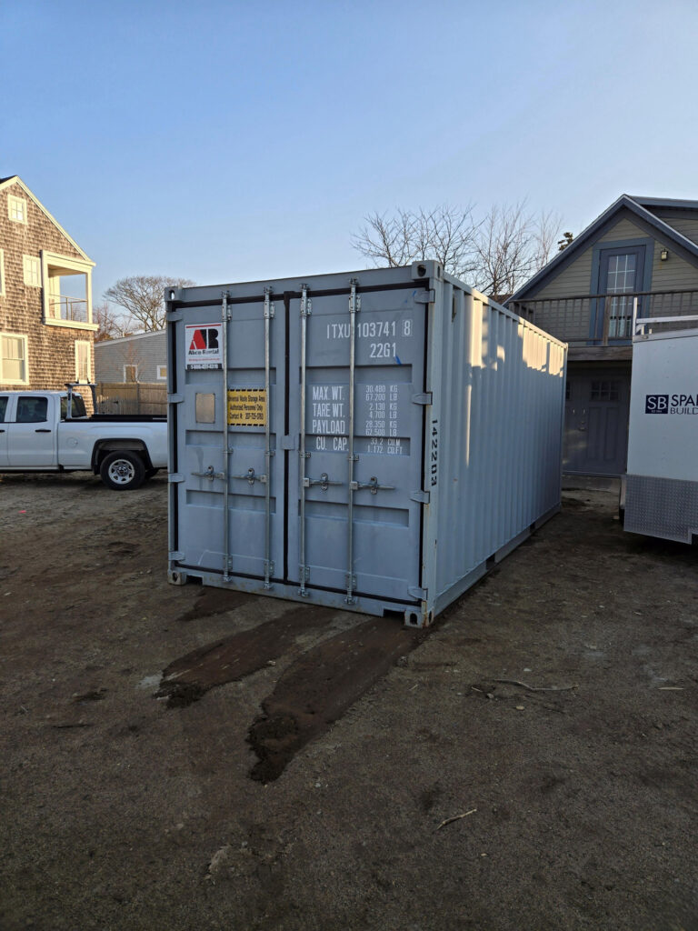 Gray shipping/storage container in a dirt lot with a white pickup truck and houses in the background under a clear blue sky.
