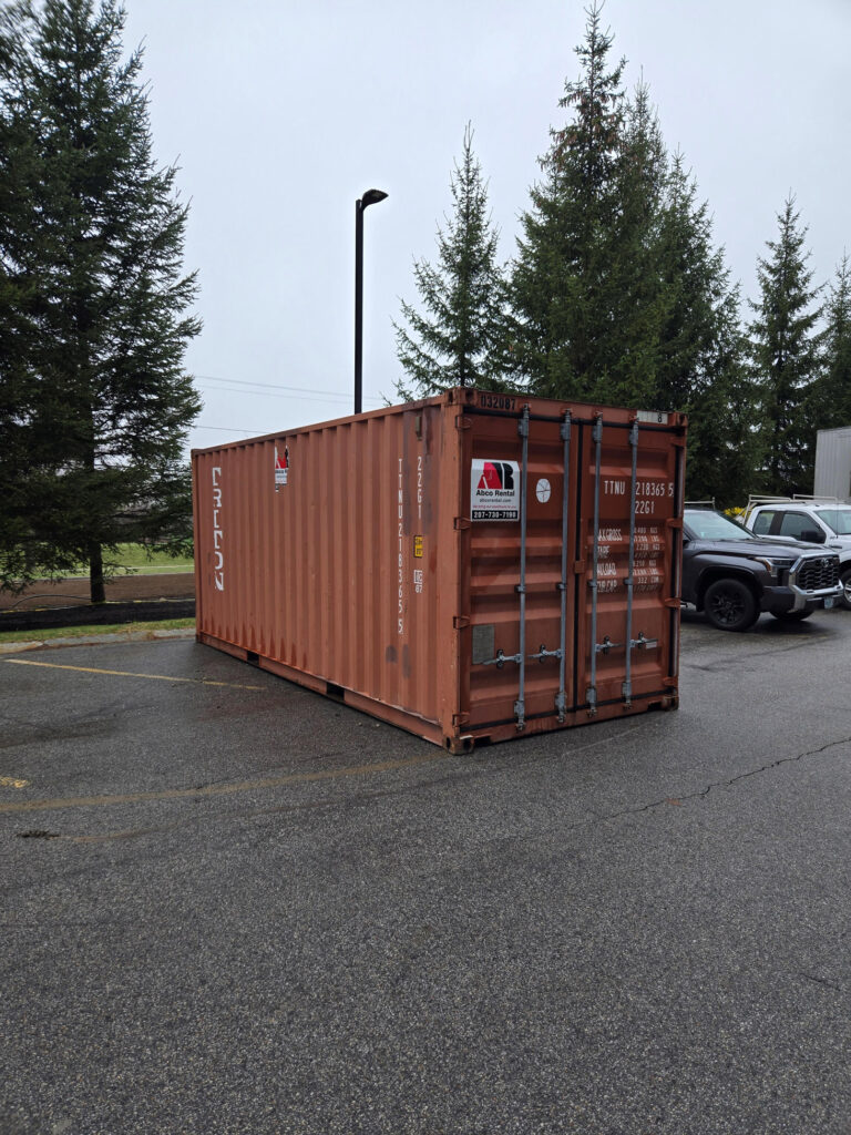 Orange shipping container in a parking lot with tall evergreen trees behind it and a streetlight nearby.