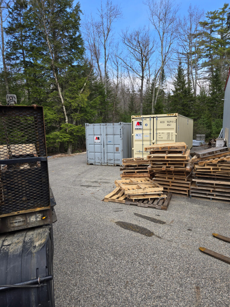 Industrial yard with stacked wooden pallets and metal shipping containers, a dirt-gray truck visible on the left, and trees in the background under a blue sky.