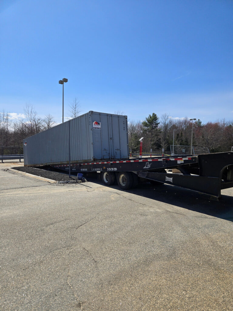 Grey shipping container on a flatbed trailer in a parking lot under a clear blue sky.